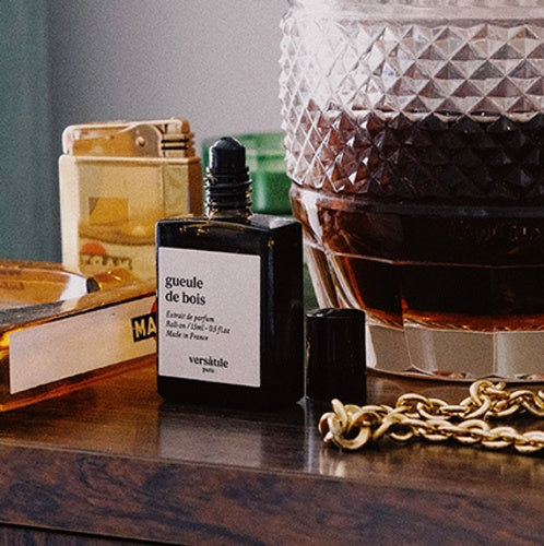 Perfume bottle labeled 'gueule de bois' on a wooden surface with a glass container and gold chain in the background.