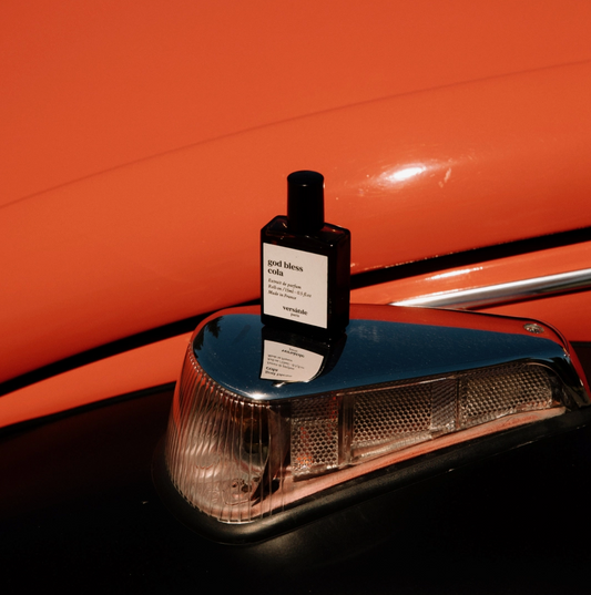 A brown glass bottle of Versatile Paris God Bless Cola perfume sits on a metallic object, in front of an orangey-red metallic curved background, which looks like it might be a car but we can't tell