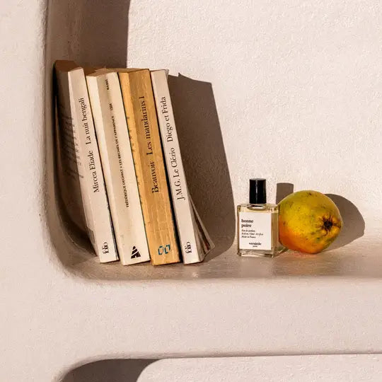 Stack of books and a bottle of perfume on a shelf with an apple beside them.