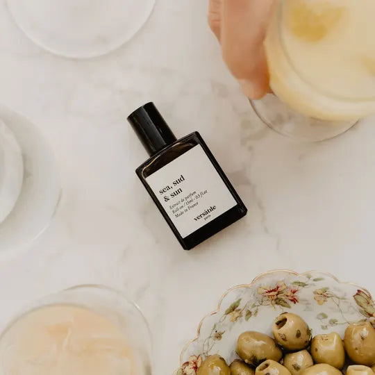 Perfume bottle labeled 'sea salt & sun' on a marble surface with olives and glasses.