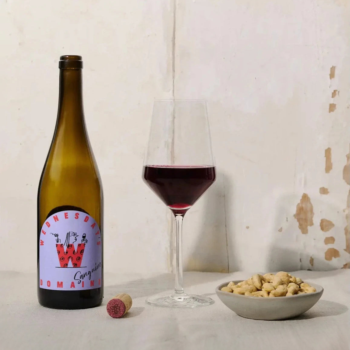 A bottle of red with with a red cap and a purple and red label stands next to a clear glass filled with wine. Some nuts sit in a white beside them.