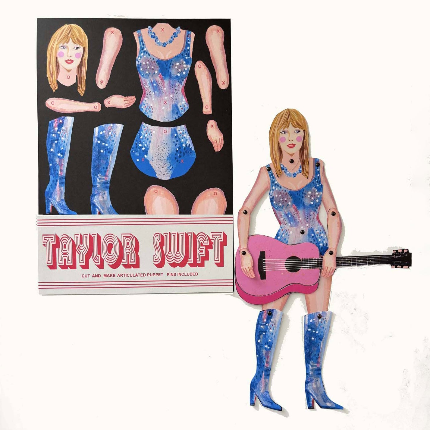 A crafted paper doll of Taylor Swift wearing a blue dress and holding a pink guitar, with a flat packaging design featuring the same image.