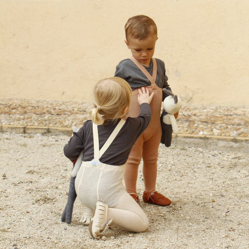 A child in a grey tshirt stands holding a soft toy in one hand stands while another child wearing cream tights and a grey t-shirt sits on one knee facing him