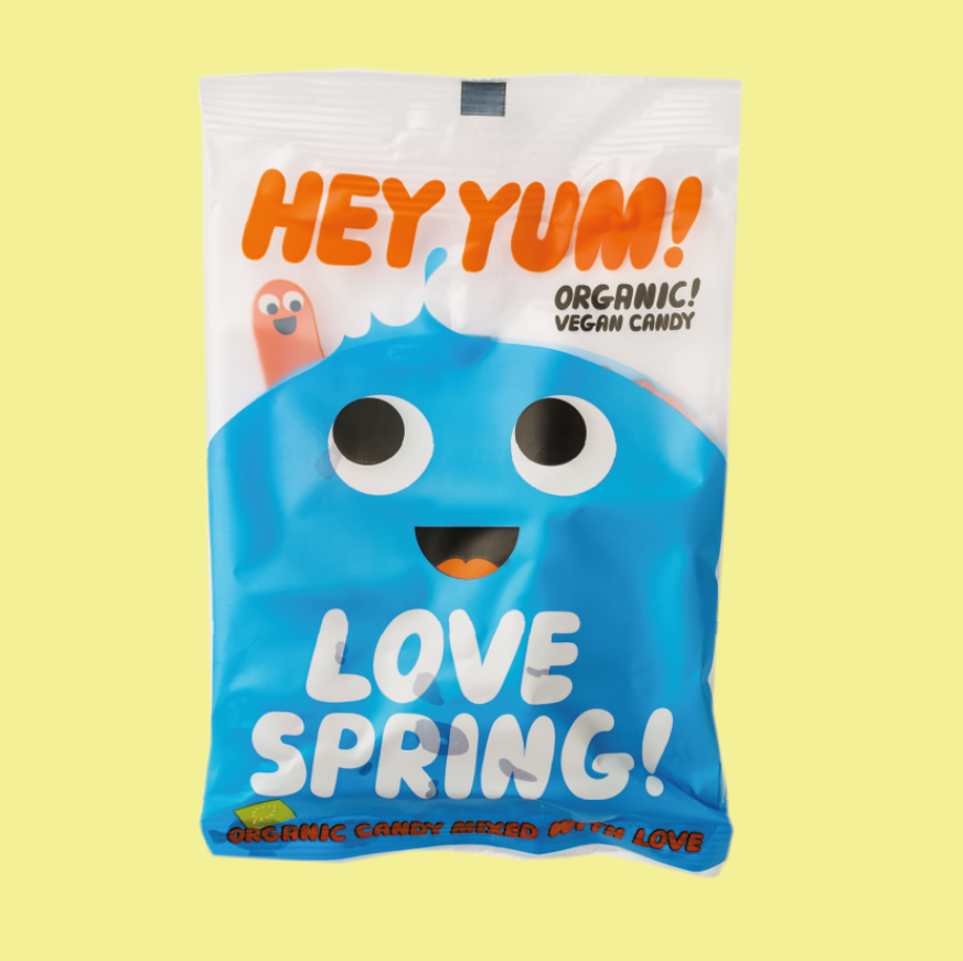 A package of HEY YUM! Love Spring, Vegan Organic Fruit Gums, with a graphic design resembling a smiling fruit character, and text stating 'LOVE SPRING!' against a blue background.
