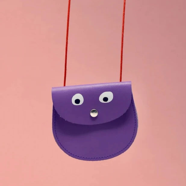 An oval shaped lilac small pocket purse with red thin rope as the bag strap hangs against a pink background. Printed onto the bag are two white googly eyes with black pupils and a small silver press button which acts as the nose and opens/closes the purse.