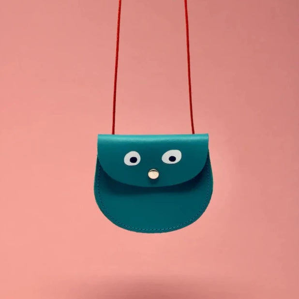 An oval shaped turquoise small pocket purse with red thin rope as the bag strap hangs against a pink background. Printed onto the bag are two white googly eyes with black pupils and a small silver press button which acts as the nose and opens/closes the purse.