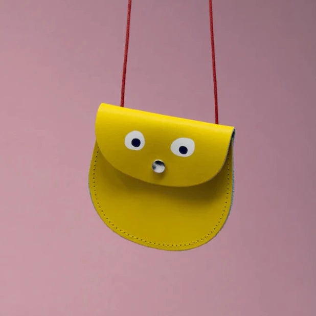 An oval shaped yellow small pocket purse with red thin rope as the bag strap hangs against a pink background. Printed onto the bag are two white googly eyes with black pupils and a small silver press button which acts as the nose and opens/closes the purse.