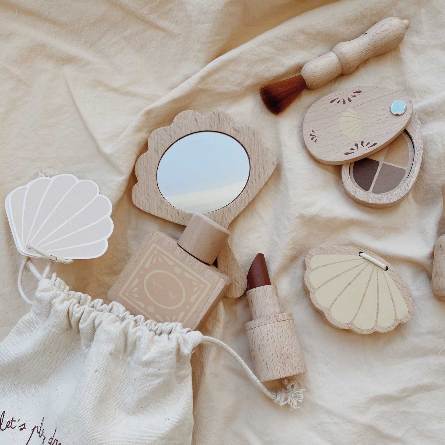 A wooden beauty play set by Konges Sløjd displayed on a soft beige fabric, featuring a shell-shaped mirror, makeup brush, lipstick, compact, and perfume bottle.