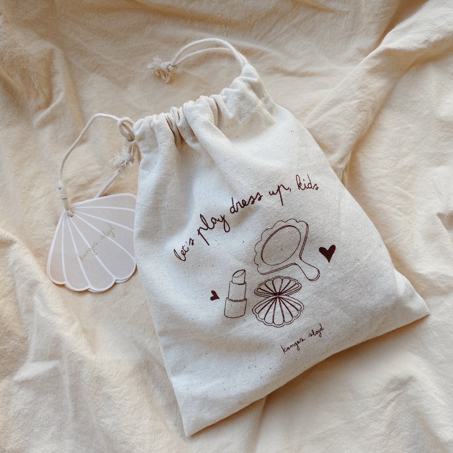 A drawstring cotton bag with "Let’s play dress up kids" printed on it, placed on a soft beige fabric, containing Konges Sløjd's wooden beauty set.