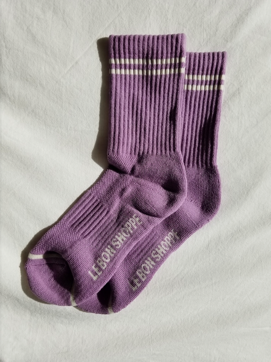 A pair of grape purple ribbed crew socks with white stripe details stacked slightly on top of each other on a white fabric background, highlighting the soft texture and branding on the sole.