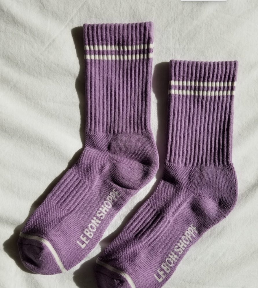 A pair of grape purple ribbed crew socks with white stripe details laid flat on a white fabric background, showing the full design, branding on the sole, and cushioned heel and toe.