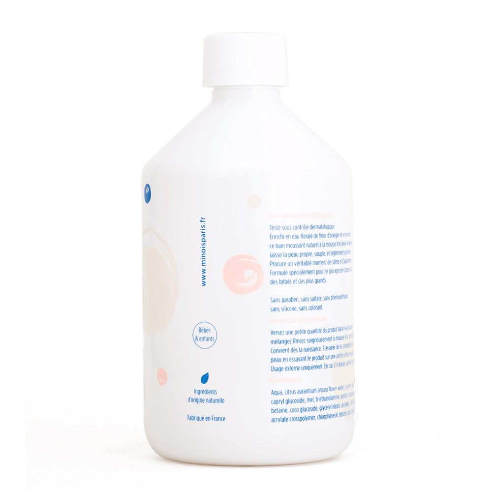 The side of a white bottle of bubble bath, with a white screw on lid. In blue there is a long list of ingredients of the product and other product information which is in French. Down the side of the bottle, it has the website of the company, that being minois and also more product information. There are also some pink and beige decorative spots. This is against a white background.