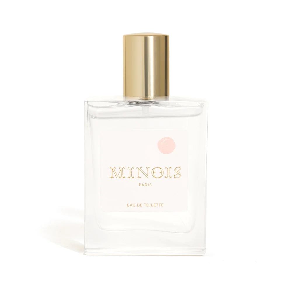 A clear square bottle of perfume with a gold lid sits in front of a white background. The bottle has a white label that says Minois in gold with a pink spot above it. Beneath Minois, also in gold it says Paris and beneath that, again in gold, it says Eau De Toilette.