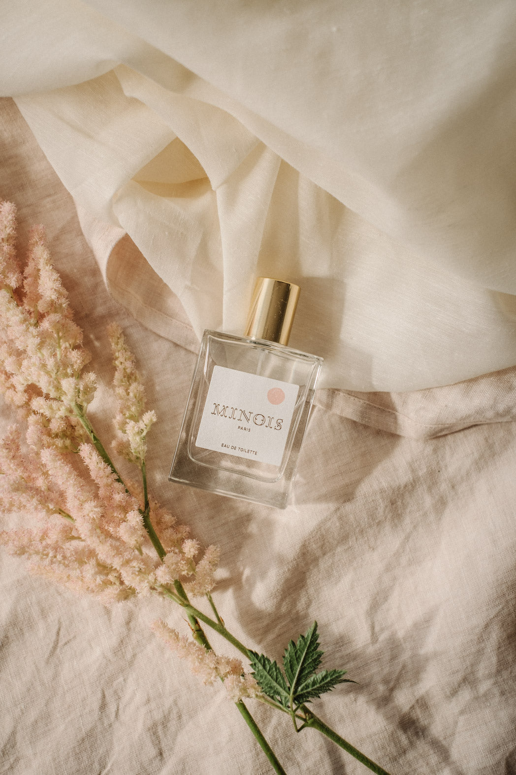 A clear square bottle of perfume with a gold lid lays on top of pink and cream sheets, next to pink feathered flowers that have a green stem. The bottle has a white label that says Minois in gold with a pink spot above it. Beneath Minois, also in gold it says Paris and beneath that, again in gold, it says Eau De Toilette.