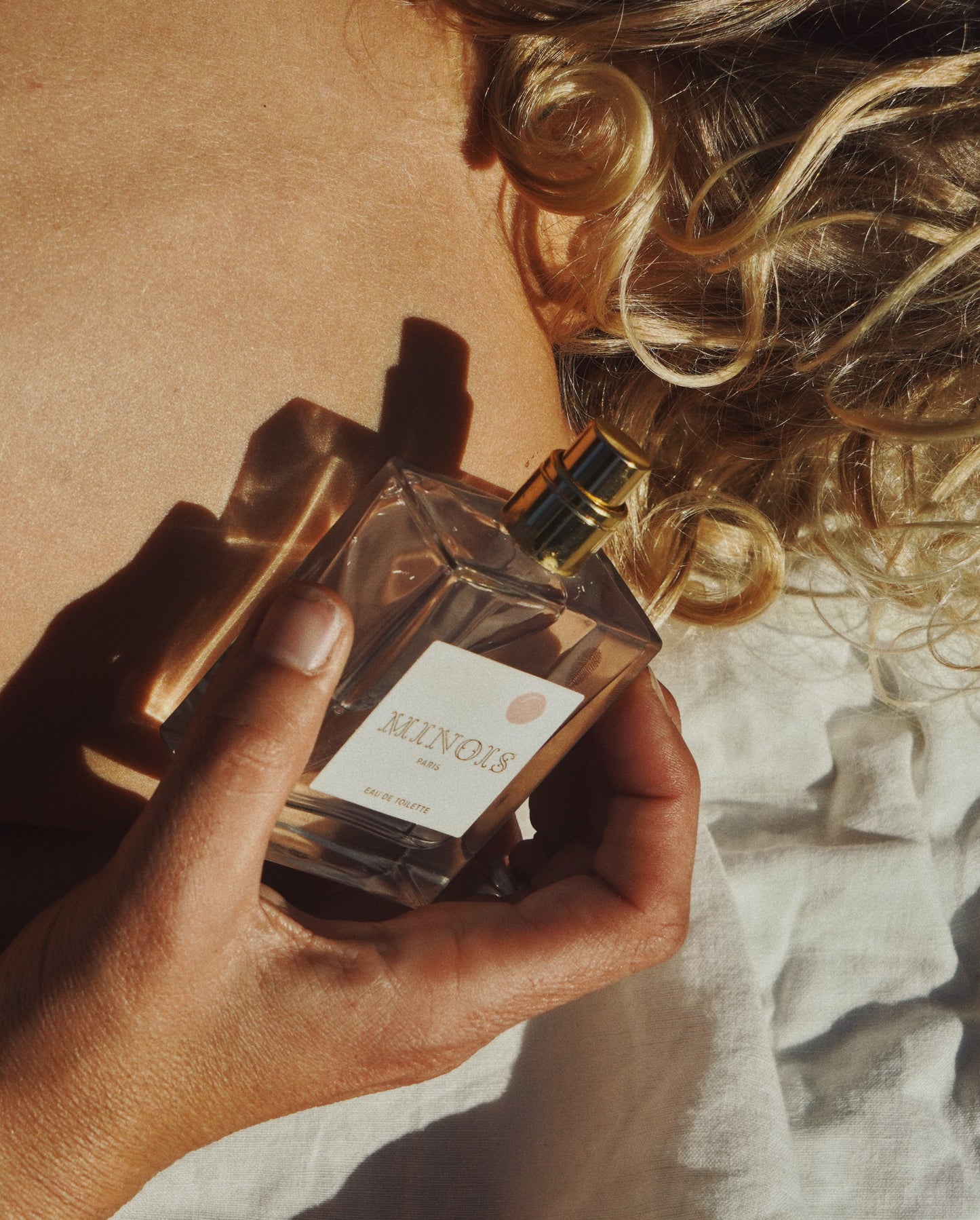 A clear square bottle of perfume with a gold lid is held up next to what appears to besomebodys back with the shadow of the bottle reflecting off their skin . The bottle has a white label that says Minois in gold with a pink spot above it. Beneath Minois, also in gold it says Paris and beneath that, again in gold, it says Eau De Toilette.