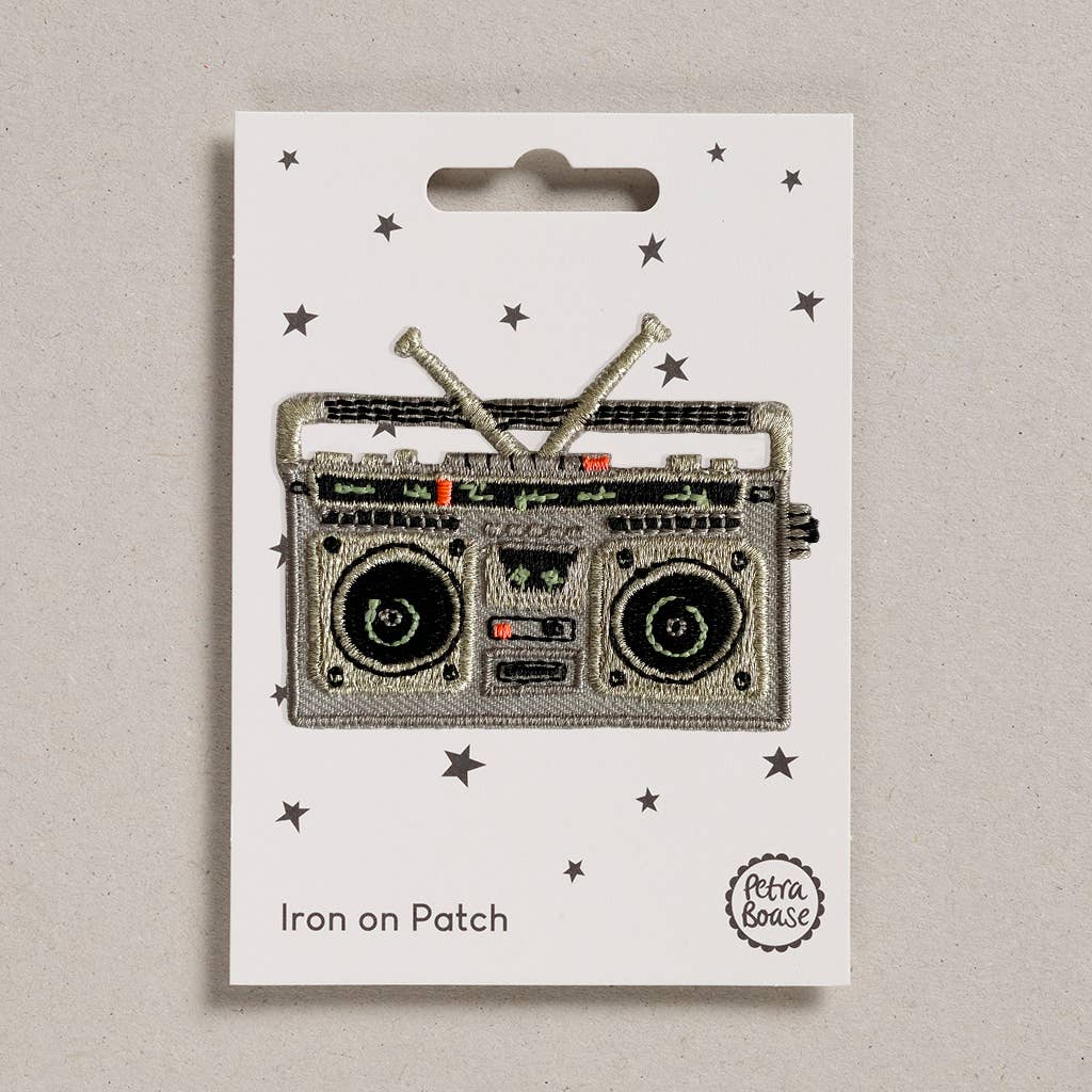 An embroidered iron-on patch shaped like a retro silver boom box, featuring detailed speakers, cassette deck, dials, and crossed antennae, with metallic silver and black stitching. Packaged on a cream backing card with black stars and Petra Boase branding.
