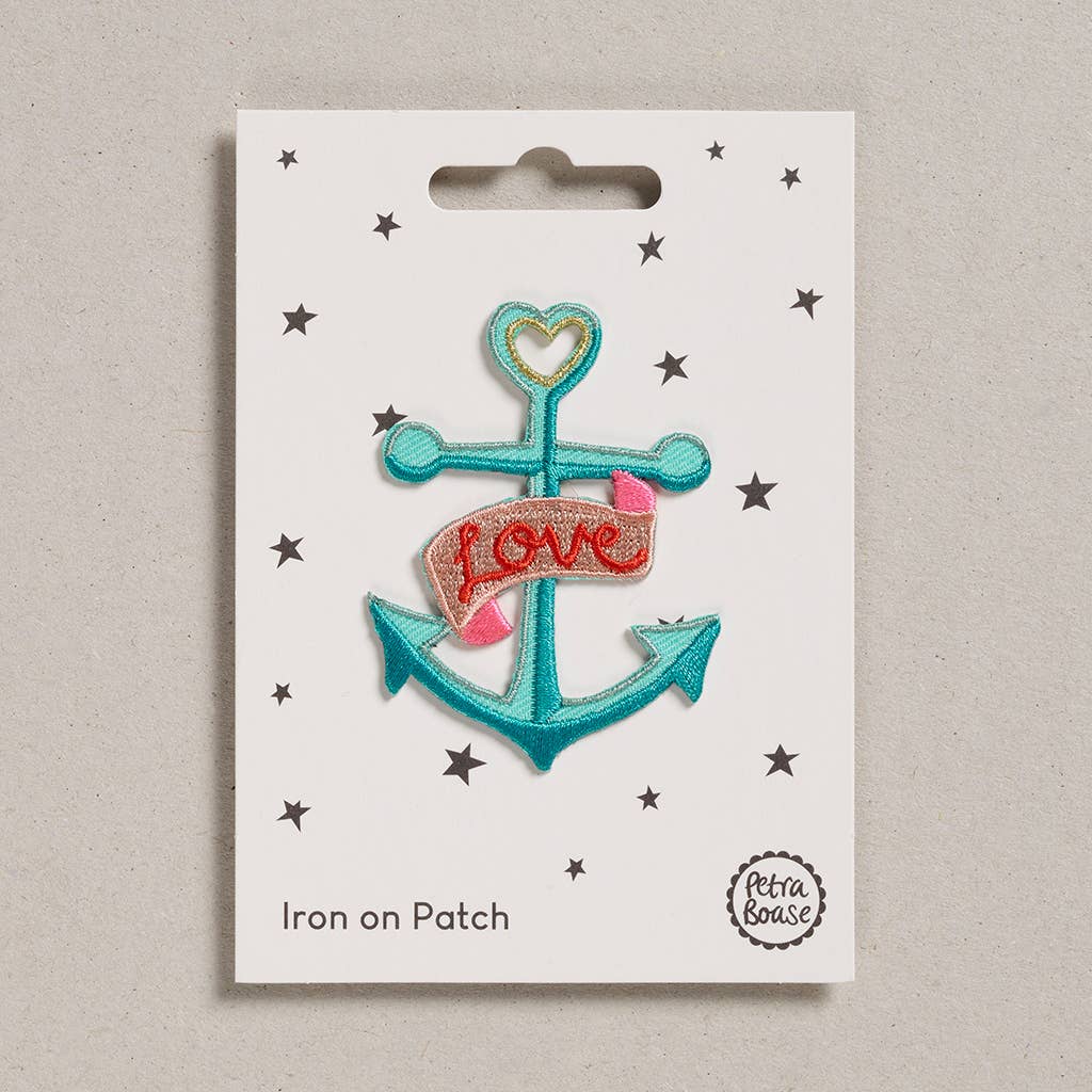 An embroidered iron-on patch featuring a vintage-style turquoise anchor with a gold heart at the top and a flowing pink banner reading "Love" in red embroidery. Packaged on a cream backing card with black stars and Petra Boase branding.