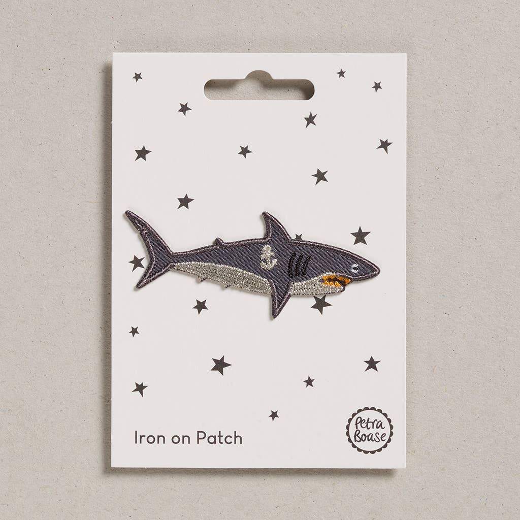 an embroidered dark and light grey shark. The gills are black in colour. He has a pink orange. He has a silver anchor on his body just by his gills.The background is white with black stars