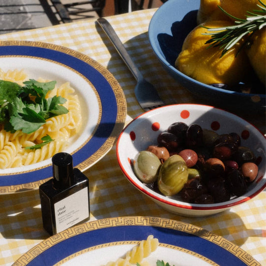 An image of a bottle of perfume, the scent being rital date, on a white and yellow checkered tablecloth, surrounded by a white bowl with red spots containing green and purple olives, a bowl with a blue trim and brown pattern surrounding it full of pasta with basil on top of it and a blue bowl with lemons and rosemary inside of it.