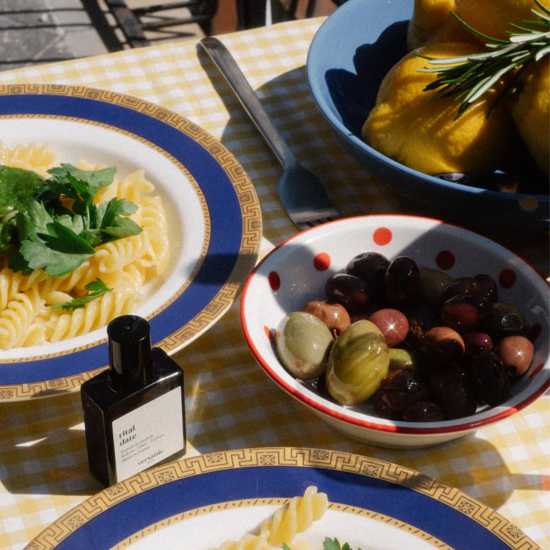 An image of a bottle of perfume, the scent being rital date, on a white and yellow checkered tablecloth, surrounded by a white bowl with red spots containing green and purple olives, a bowl with a blue trim and brown pattern surrounding it full of pasta with basil on top of it and a blue bowl with lemons and rosemary inside of it.