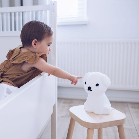 A child inside a crib reaching to touch a dog shaped Snuffy First Light Lamp placed on a wooden stool next to the white crib in white bright lit room.