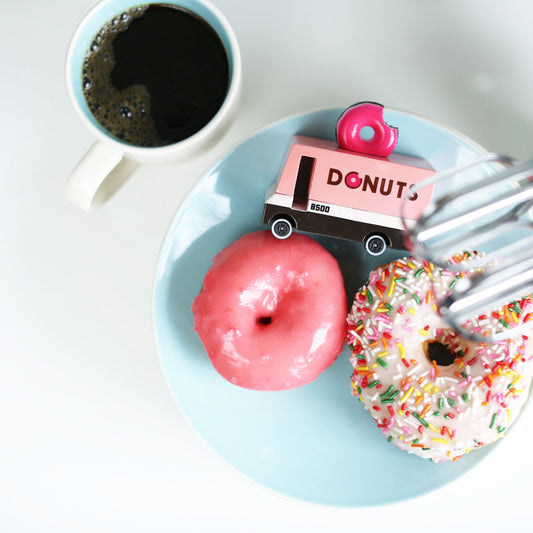 A toy donut van with a pink topper placed on a plate with two actual donuts, alongside a cup of coffee.