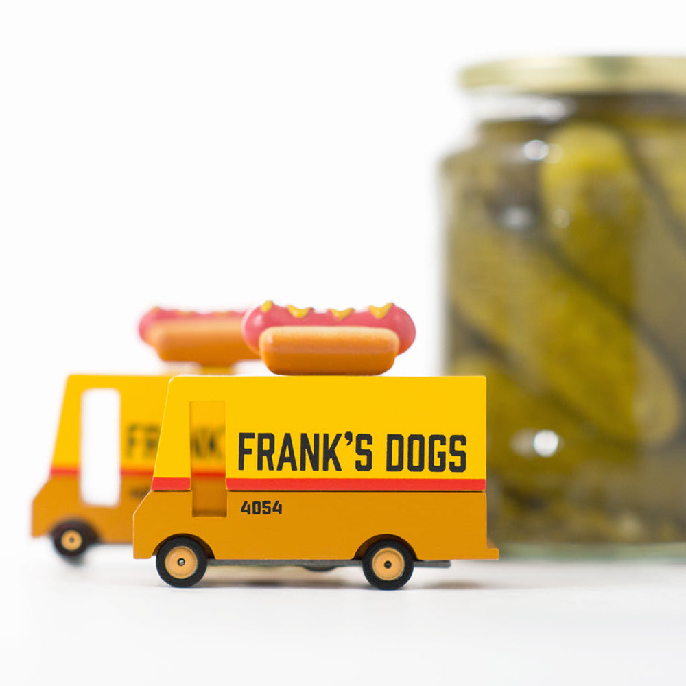 A toy hotdog van with a mustard-yellow top and a red hotdog on the roof, labeled 'FRANK'S DOGS', in front of a jar of pickles.