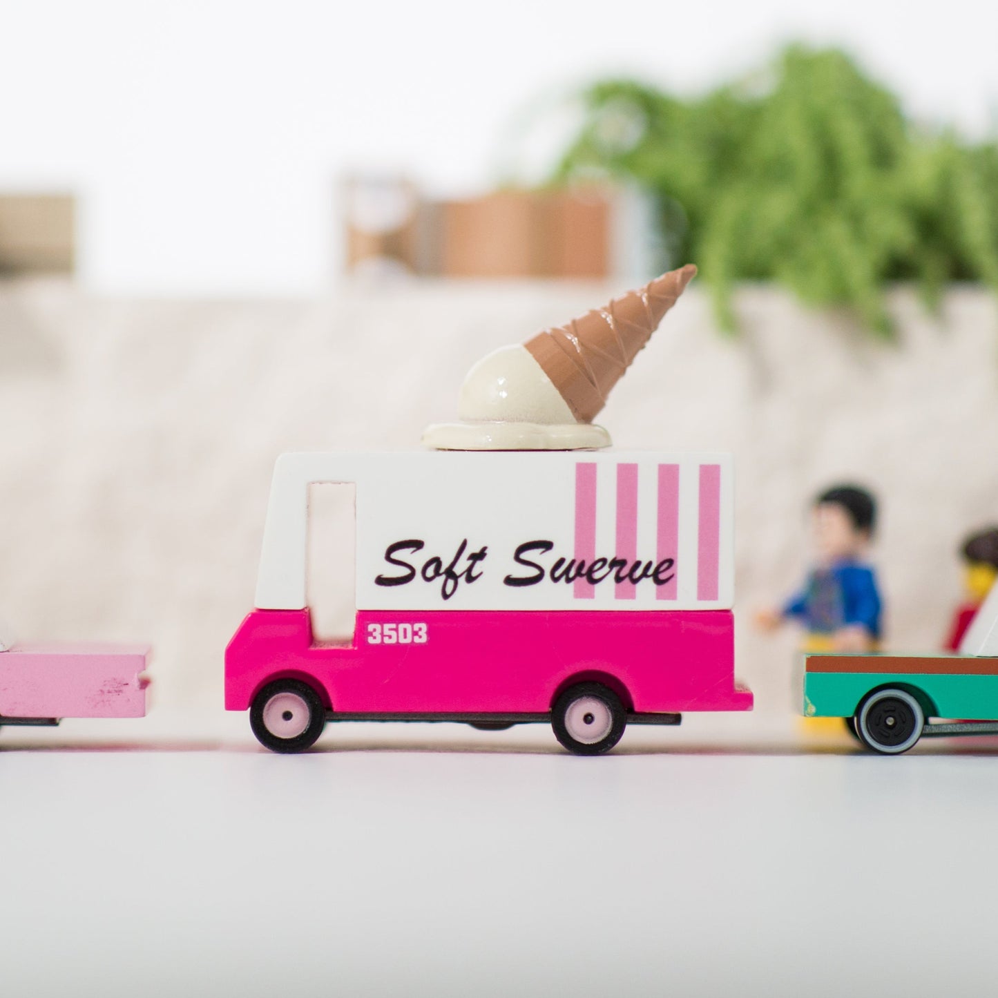 A toy ice cream van with a pink base and a white ice cream cone on top, labeled 'Soft Serve'. It is part of a playset that includes other toy cars.