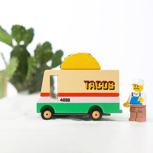 A wooden toy taco truck with a 'TACOS' sign on top, painted in multiple colors, including orange, green, and yellow. A small figurine of a chef is standing next to the truck.