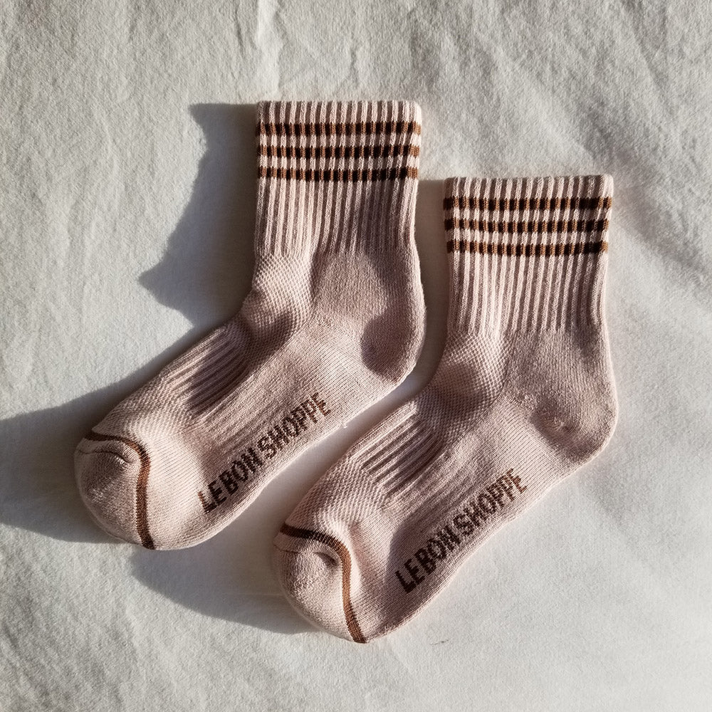 A pair of Bellini pink ribbed crew socks with brown stripes, laid flat on a white fabric background, captured in soft natural light, emphasizing the texture and embroidered branding on the sole.