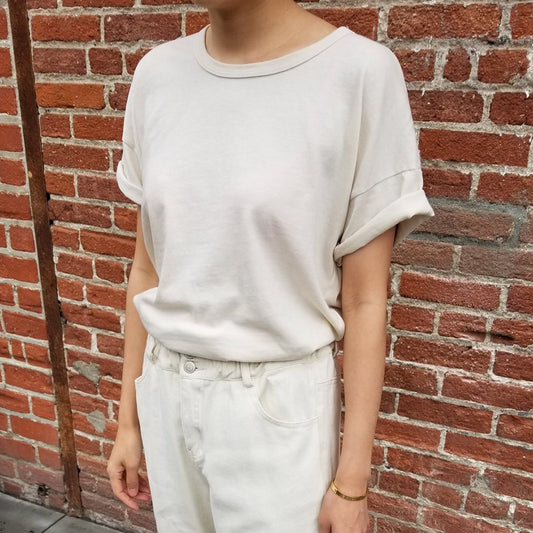 A person wearing a beige, round-neck, dropped shoulder t-shirt with rolled-up sleeves and a shirttail hem, paired with light-colored pants, standing in front of a brick wall.