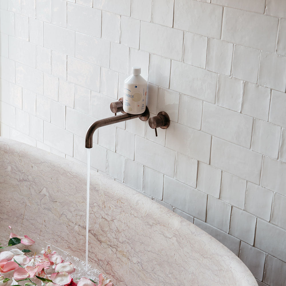 A white bottle of minois bubble bath sits on top of a rusted gold running tap with two rusted gold handles either side against an off-white tiled wall. This is above a rose gold marble bath which has pink flower petals inside it .