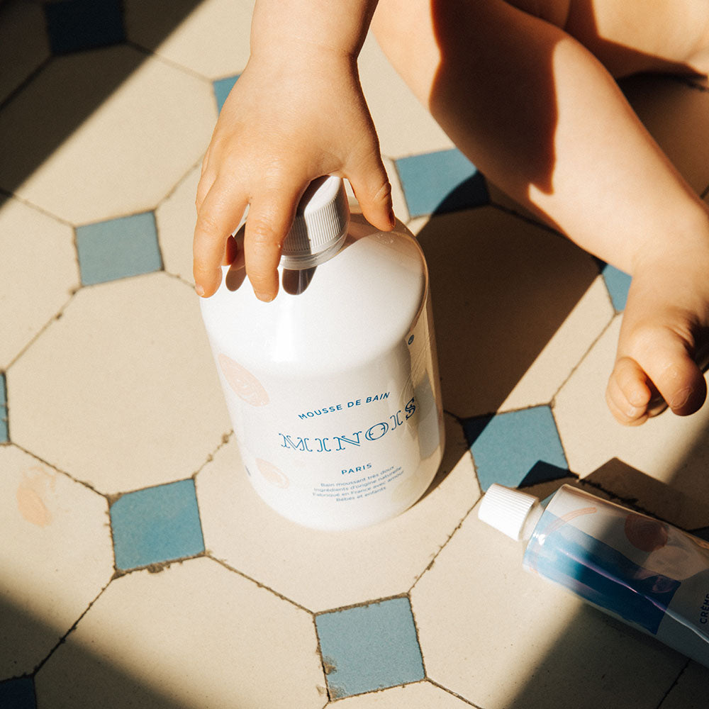 A white bottle of minois bubble bath is being held by the cap by what appears to be a children's hand. The bottle is on a white tiled floor which has blue diamond tiles around each white square tile. The child appears to be siting down as their foot and leg are in the image. There is also a tube of what appears to be cream also on the floor.