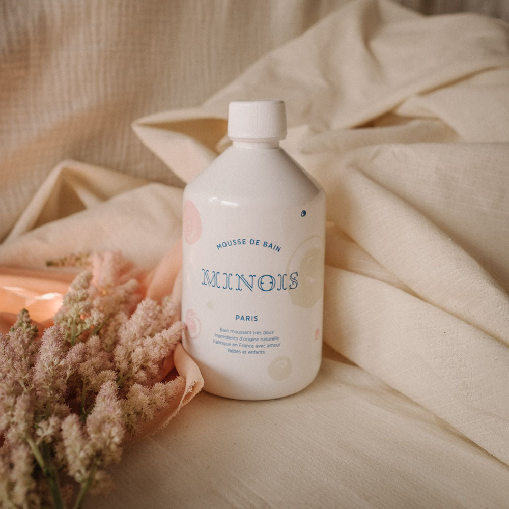 A white bottle of bubble bath, with a white screw on lid, that in blue says Mousse De Bain at the top which means bath foam, beneath that it says Minois, beneath that it says Paris, sits on top of light pink/beige sheets next to pink feathery flowers. Beneath Paris there is smaller text in French which describes the ingredients of the product, where it was made and its suitability for children. The bottle itself is decorated with pink and beige spots.