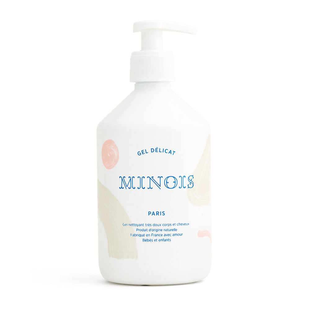 A white bottle of washing gel, with a white pump lid that. In blue at the top of the bottle it says Gel Delicat at the top, beneath that it says Minois, beneath that it says Paris. Beneath Paris there is smaller text in French which describes the ingredients of the product, where it was made and its suitability for children. The bottle itself is decorated with pink spots and beige patterns. This is against a white background.