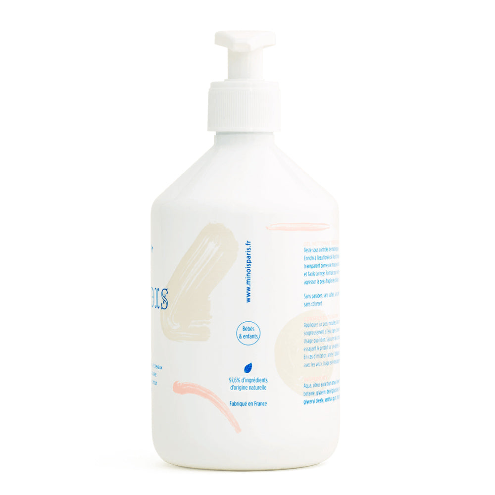 The side of a white bottle of washing gel, with a white pump lid. In blue there is a long list of ingredients of the product and other product information which is in French. Down the side of the bottle, it has the website of the company, that being minois and also more product information. There are also some pink and beige decorative lines. This is against a white background.