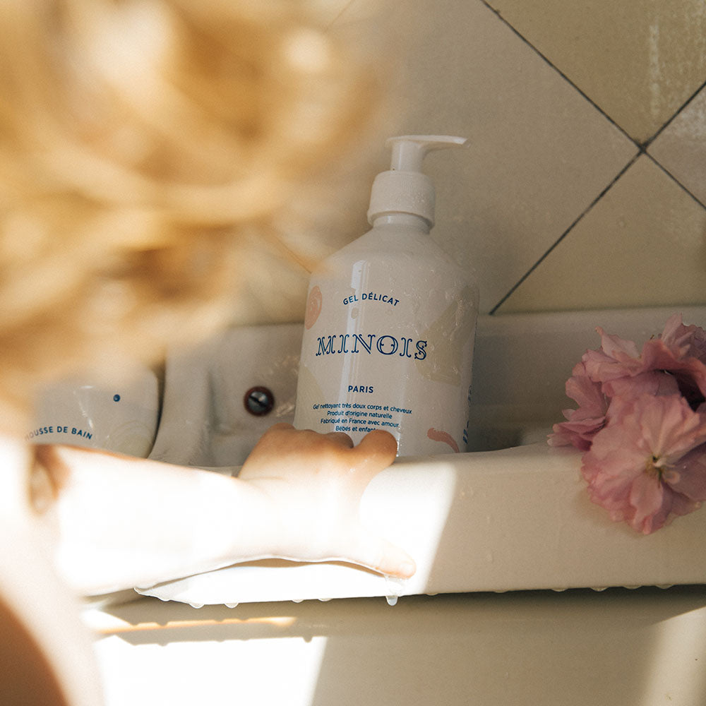A white bottle of washing gel, with a white pump lid that sits inside of what appears to be a white ledge above a bath next to pink flowers, being held onto by a child. In blue at the top of the bottle it says Gel Delicat at the top, beneath that it says Minois, beneath that it says Paris. Beneath Paris there is smaller text in French which describes the ingredients of the product, where it was made and its suitability for children. The bottle itself is decorated with pink spots and beige patterns.