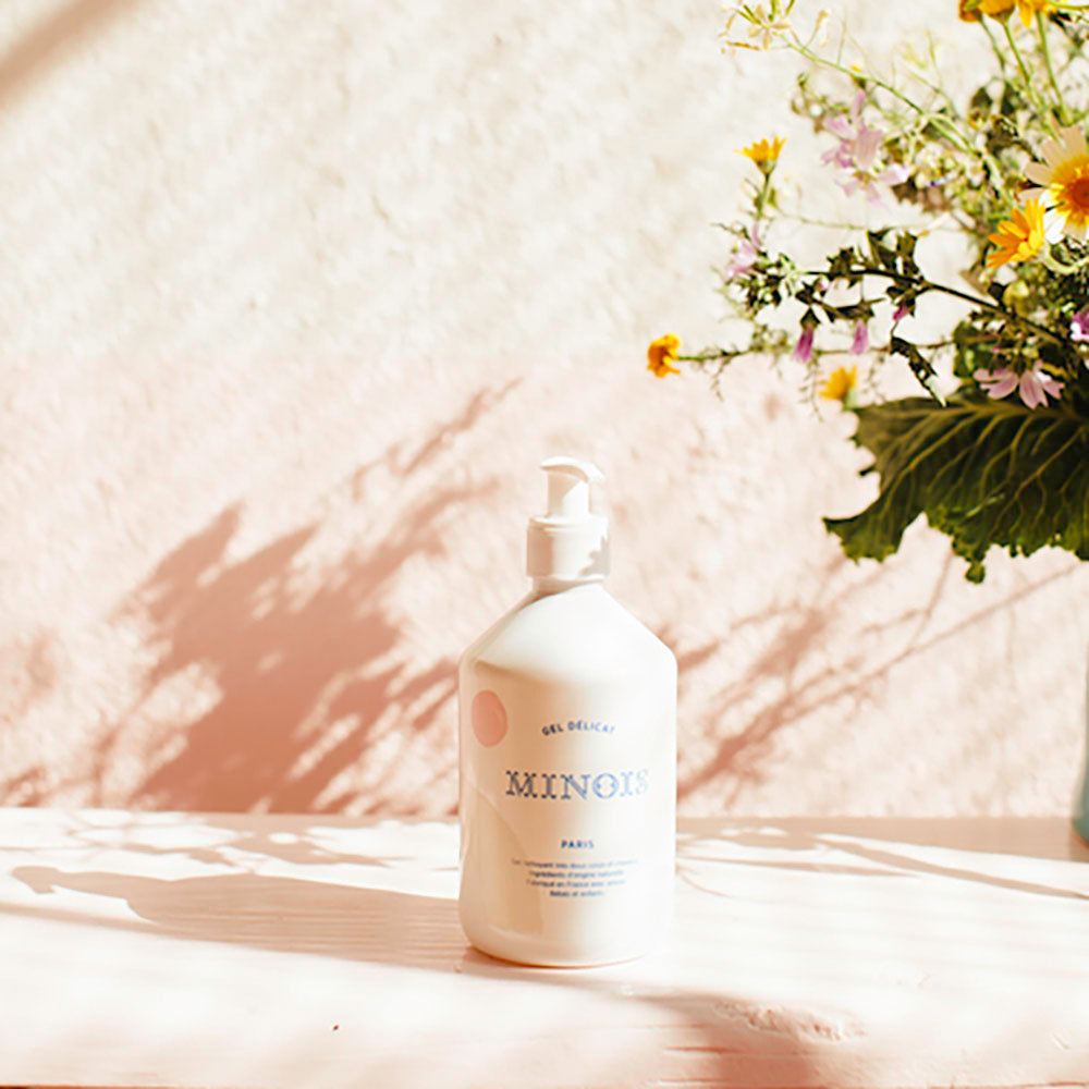 A bottle of minois gentle washing gel sits on what appears to be a cream/pink wooden table or bench in front of a pink wall. Next to the bottle thee is the top of bunch of pink, yellow, green and cream flowers, with the shadow of them reflecting off the wall In blue at the top of the bottle it says Gel Delicat at the top, beneath that it says Minois, beneath that it says Paris. Beneath Paris there is smaller text in French which describes the ingredients, where its made and its child suitability.