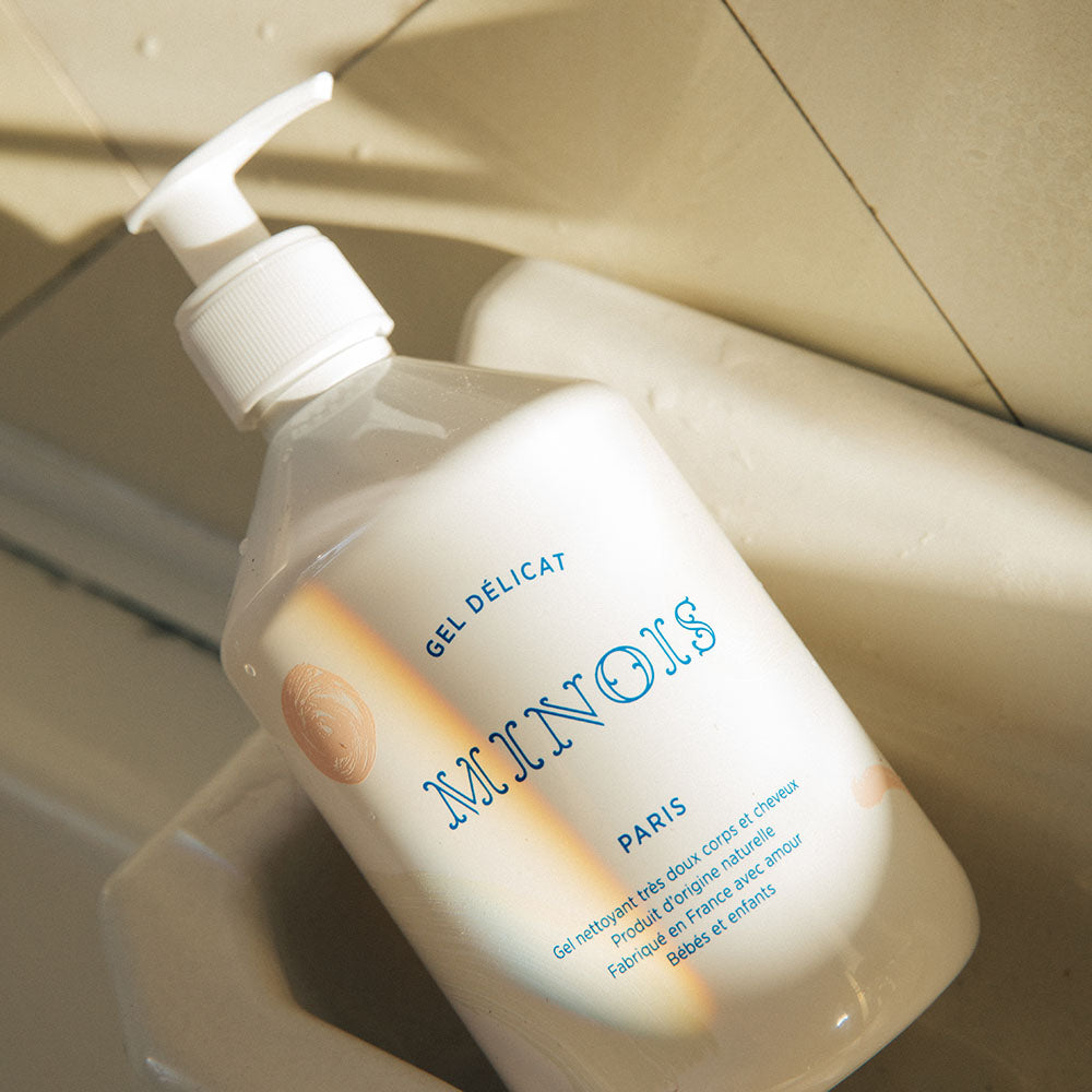 An overhead shot of a white bottle of washing gel, with a white pump lid that sits inside of what appears to be a white ledge above a bath. In blue at the top of the bottle it says Gel Delicat at the top, beneath that it says Minois, beneath that it says Paris. Beneath Paris there is smaller text in French which describes the ingredients of the product, where it was made and its suitability for children. The bottle itself is decorated with pink spots and beige patterns.
