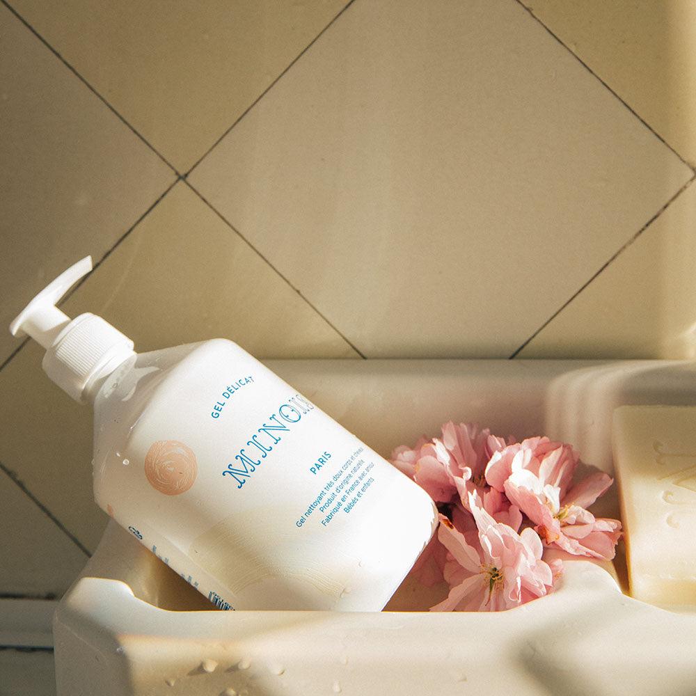 A white bottle of washing gel, with a white pump lid that sits inside of what appears to be a white ledge above a bath next to pink flowers and a bar of soap. In blue at the top of the bottle it says Gel Delicat at the top, beneath that it says Minois, beneath that it says Paris. Beneath Paris there is smaller text in French which describes the ingredients of the product, where it was made and its suitability for children. The bottle itself is decorated with pink spots and beige patterns.