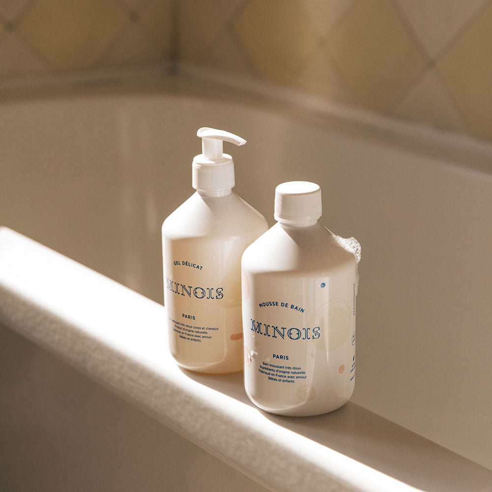 A bottle of minois bubble bath and minois washing gel sit on the edge of a white bathtub. The bottle of bubble bath has some bubbles on it and both bottles have the reflection of what appears to be light from a window.