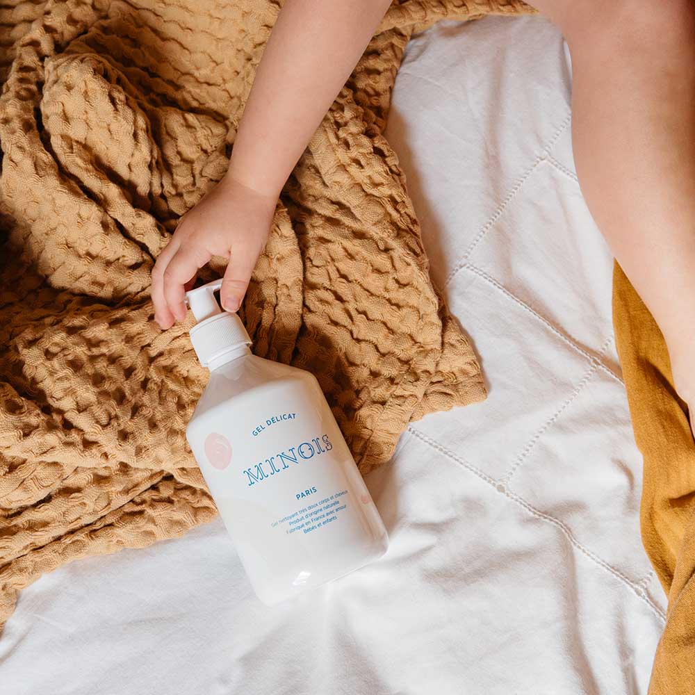 A white bottle of washing gel, with a white pump lid that sits on top of white sheets and a brown blanket, held in the hand of a child. In blue at the top of the bottle it says Gel Delicat at the top, beneath that it says Minois, beneath that it says Paris. Beneath Paris there is smaller text in French which describes the ingredients of the product, where it was made and its suitability for children. The bottle itself is decorated with pink spots and beige patterns.