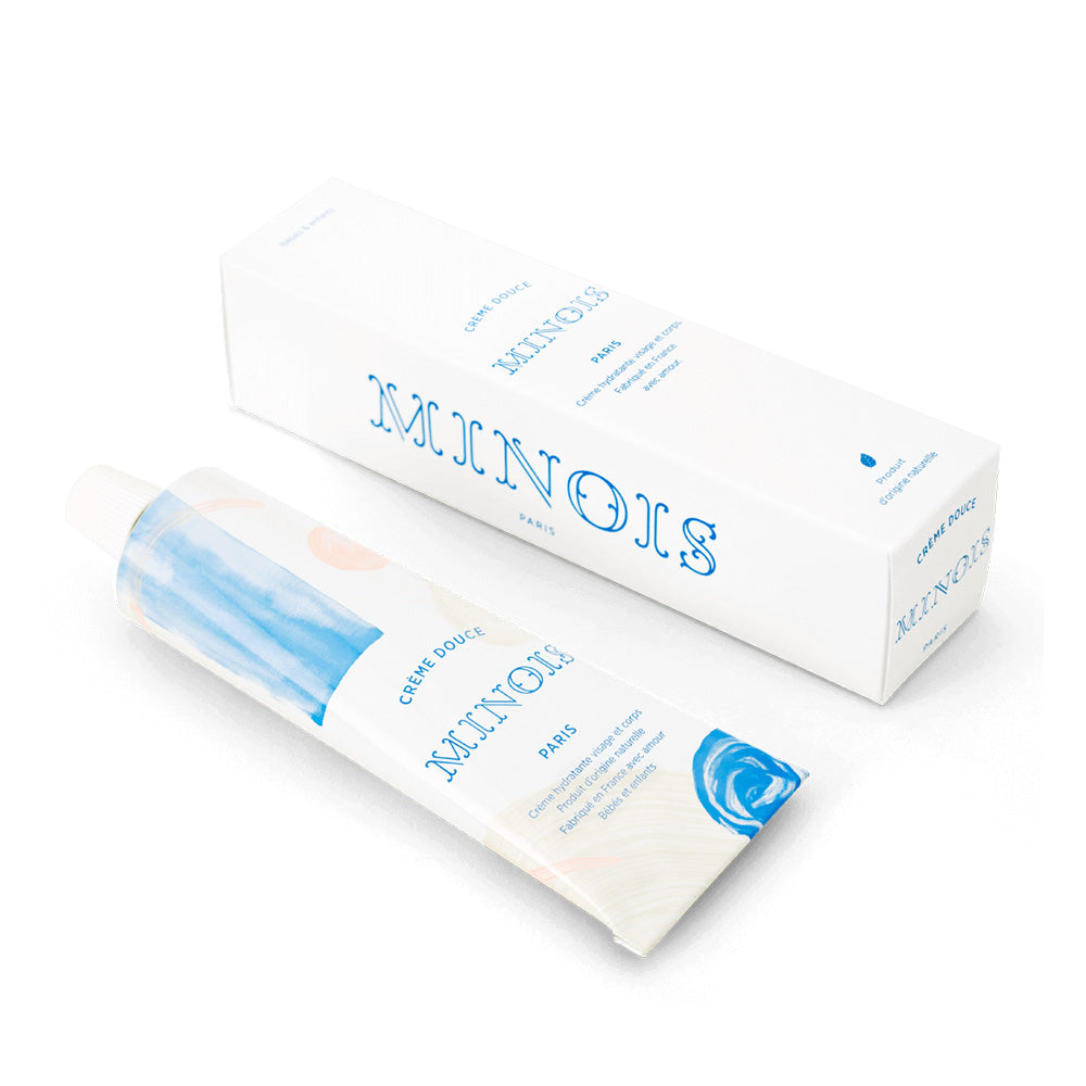 A white squeeze tube with patterns of blue and pink circles, cream and pink lines and blue squares in front of a white background lays next to the packaging for the product which is white, says Minois along the side of the packaging and has the same product information on the front of it. On the tube, in blue, it says Creme Douce, followed by Minois beneath that and Paris beneath that. Underneath Paris, still in blue in French it lists its child suitability, the natural ingredients and where it was made.