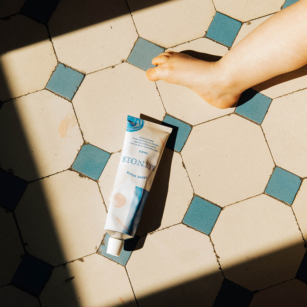A white squeeze tube with patterns of blue and pink circles, cream and pink lines and blue squares lays on top of white square bathroom floor tiles that have blue diamond square tiles next to them, next to the foot of a child. On the tube, in blue, it says Creme Douce, followed by Minois beneath that and Paris beneath that. Underneath Paris, still in blue in French it lists its child suitability, the natural ingredients and where it was made.