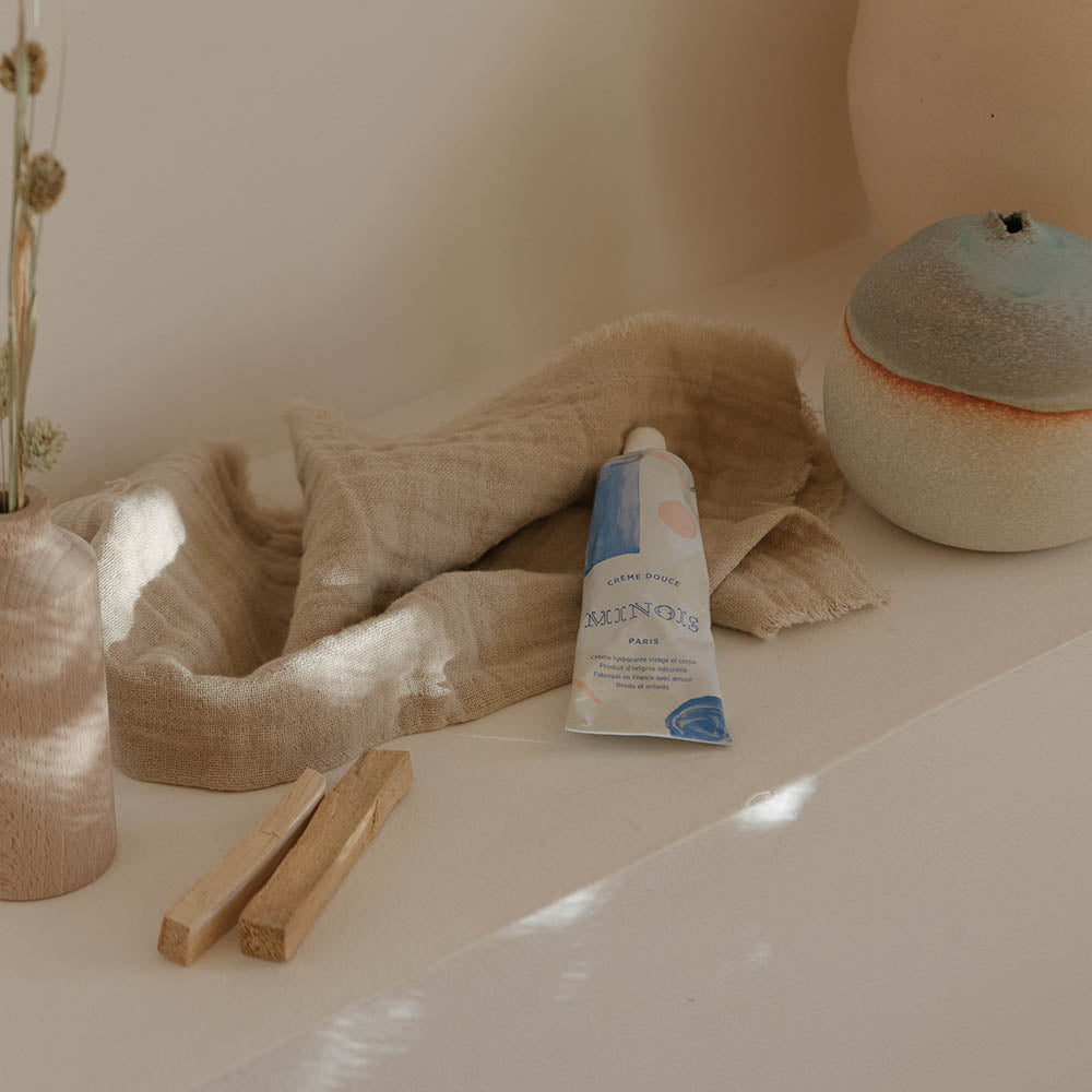 A white squeeze tube with patterns of blue and pink circles, cream and pink lines and blue squares sits on a white shelf on top of a brown cloth, next to a white ceramic with a red line and blue above it, wooden pegs and wood vase with green and brown flowers. On the tube, in blue, it says Creme Douce, followed by Minois beneath that and Paris beneath that. Underneath Paris, still in blue in French it lists its child suitability, the natural ingredients and where it was made.