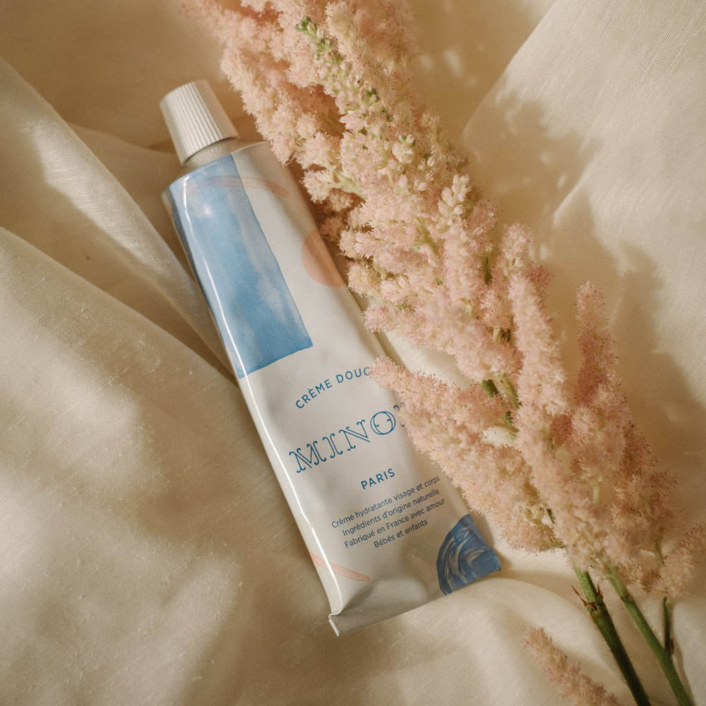 A white squeeze tube with patterns of blue and pink circles, cream and pink lines and blue squares lays on top if cream sheets next to baby pink feathery flowers that have a green stem. On the tube, in blue, it says Creme Douce, followed by Minois beneath that and Paris beneath that. Underneath Paris, still in blue in French it lists its child suitability, the natural ingredients and where it was made.