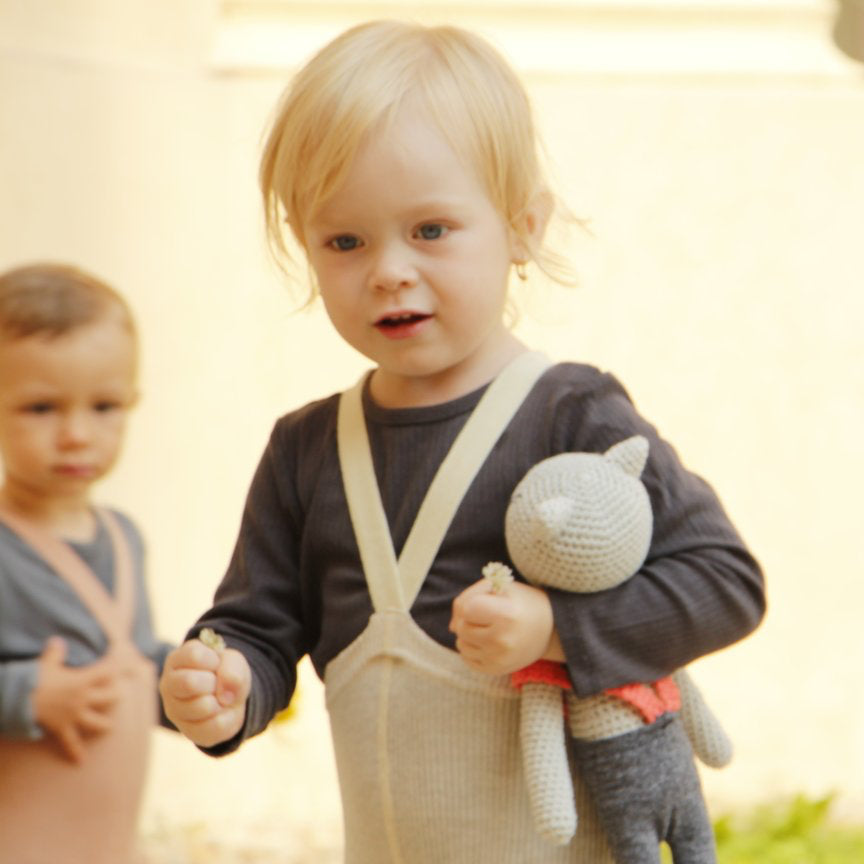 A child wearing cream tights with braces and a grey full sleeve t-shirt holds a soft toy in one hand. Another child wearing light brown tights with braces and a blue t-shirt in the background but out of focus.