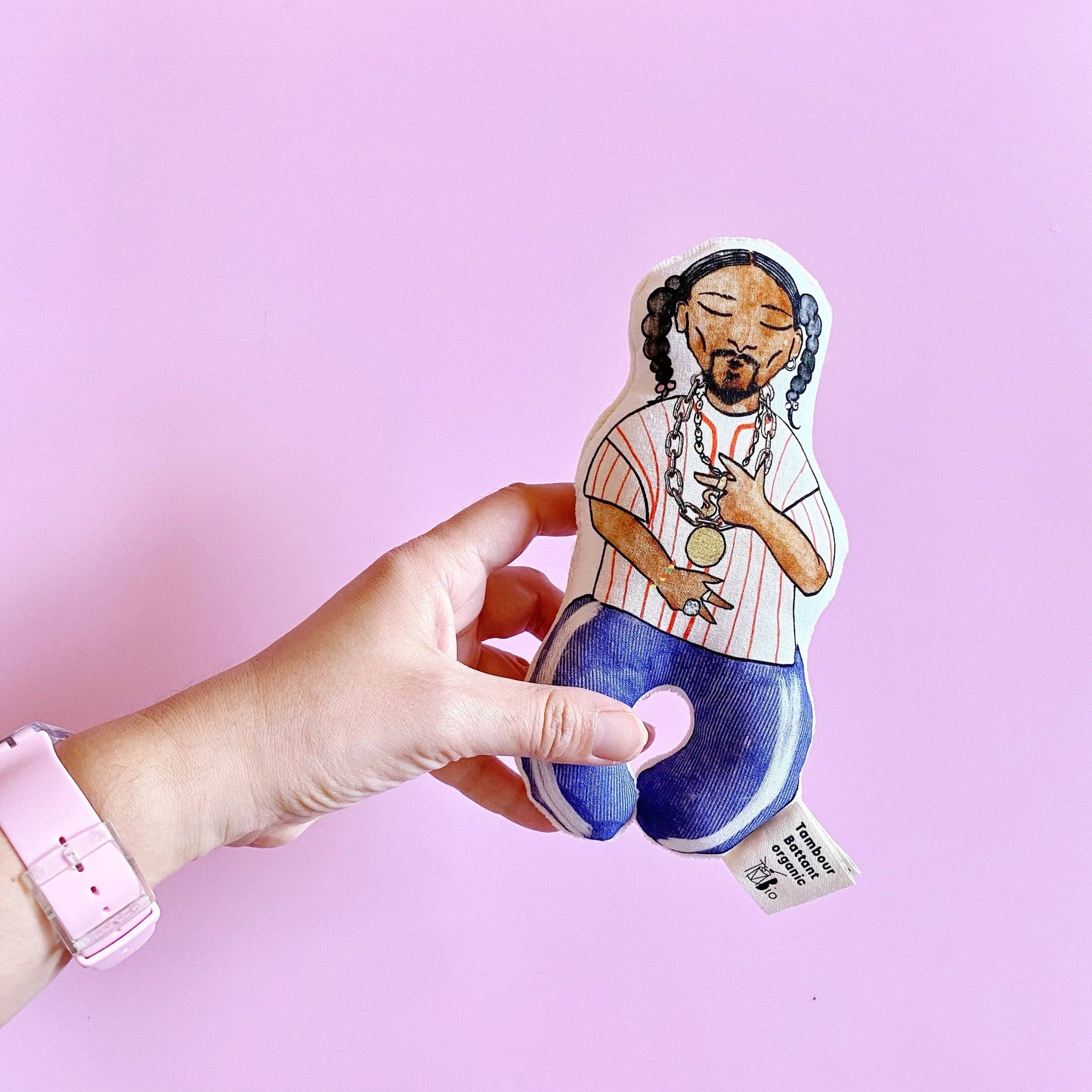 A hand holding a colorful baby rattle designed to resemble a character, with a pink background.