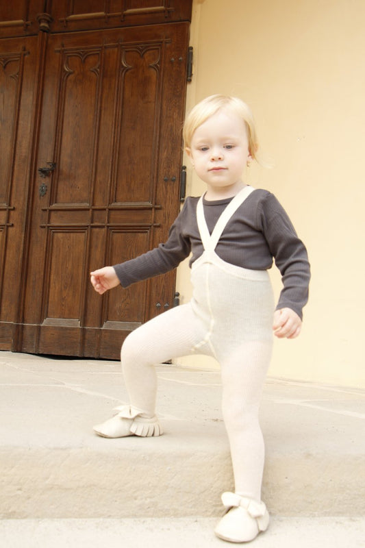 A child wearing cream tights with braces and a grey full sleeve t-shirt stands on top of a step in front of wooden doors.