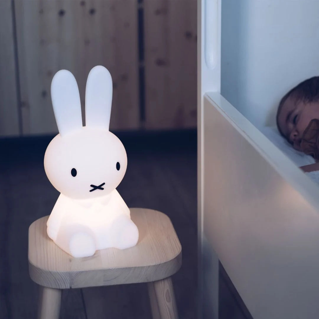 A glowing Miffy First Light Lamp sits on a wooden stool beside a sleeping baby in a dimly lit nursery.